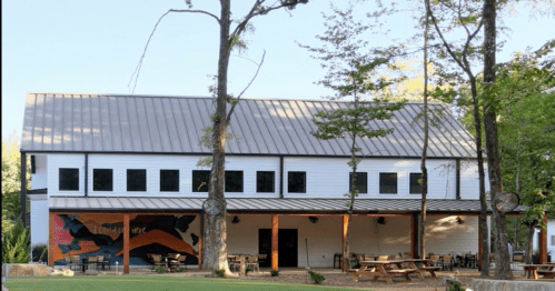 A modern building with a metal roof, large windows, and a mural, surrounded by trees and outdoor seating.