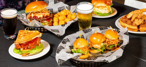 A spread of burgers, chicken and waffles, tater tots, and drinks on a table.