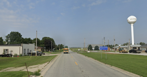 A rural road with a water tower in the background, flanked by buildings and vehicles on a clear day.
