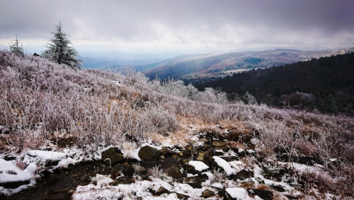A snowy landscape with frosted trees and rocky terrain, overlooking a misty valley and distant mountains.