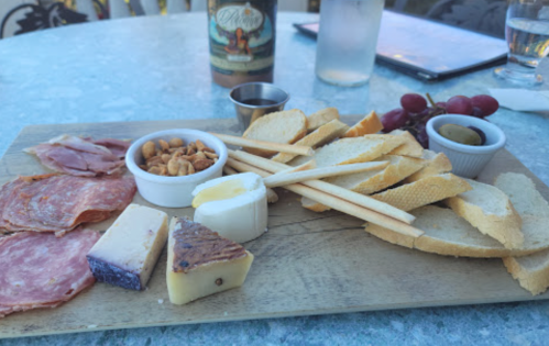 A wooden platter with assorted cheeses, cured meats, breadsticks, nuts, and grapes, set on a table.