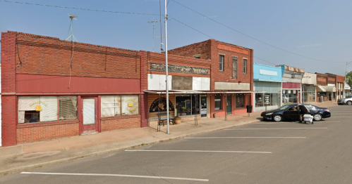 A small-town street with brick buildings, shops, and parked cars under a clear blue sky.