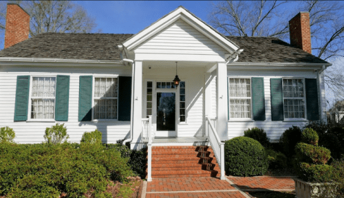 A white house with green shutters, a front porch, and brick steps, surrounded by neatly trimmed bushes and trees.