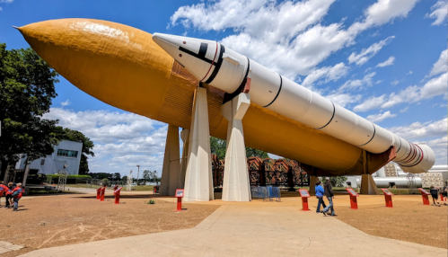 A large rocket on display outdoors, with people walking nearby and a blue sky with clouds in the background.