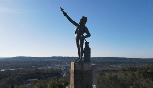 A tall statue of a man pointing upward, standing on a pedestal with a scenic landscape in the background.