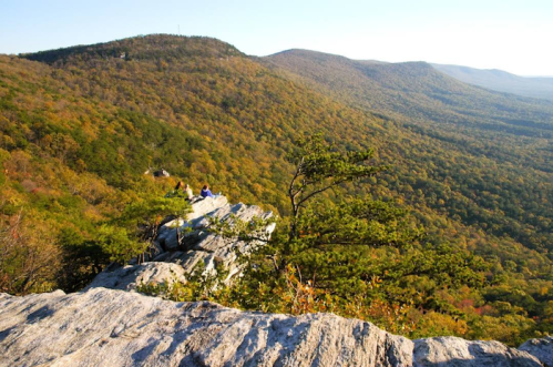 A scenic view from a rocky outcrop overlooking a valley filled with autumn-colored trees and distant mountains.