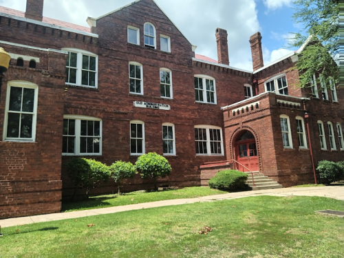 Historic red brick building with large windows and a welcoming entrance, surrounded by green grass and trees.