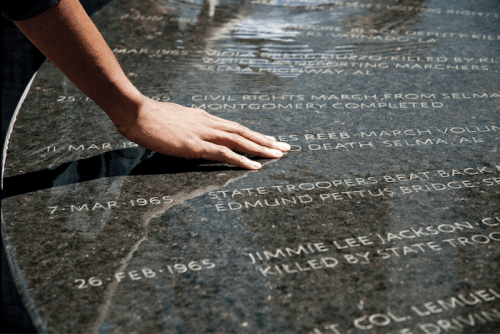 A hand rests on a reflective stone memorial inscribed with historical civil rights events and dates.