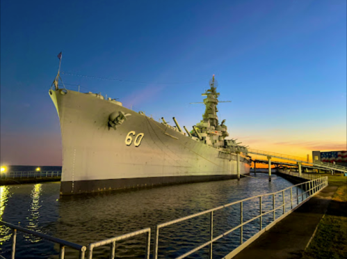 A battleship docked at sunset, with a calm water reflection and a clear sky transitioning to twilight.