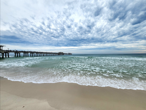 A serene beach scene with gentle waves, a pier in the distance, and a cloudy sky above.