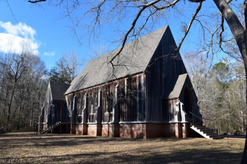 A large, rustic wooden church with gothic-style windows, surrounded by bare trees and a clear blue sky.