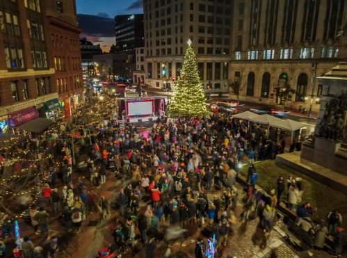 A bustling city square filled with people celebrating around a large Christmas tree and festive lights at dusk.