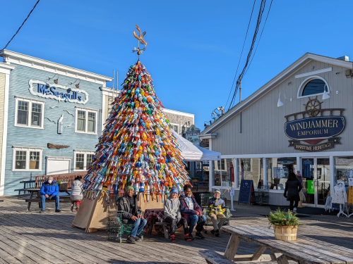 A colorful Christmas tree made of buoys stands on a boardwalk, with people sitting nearby and shops in the background.