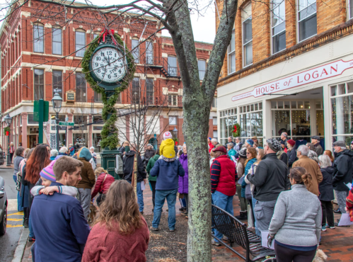A crowd gathers around a large clock in a historic town square, with shops and trees in the background.