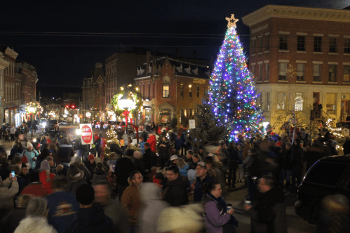 A festive street scene with a large, decorated Christmas tree and a crowd enjoying holiday lights and decorations at night.