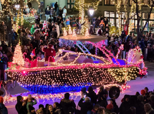 A festive boat decorated with colorful lights, surrounded by a cheering crowd at a holiday parade.