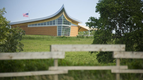 Modern building with a curved roof, set on a grassy hill, with an American flag and trees in the foreground.