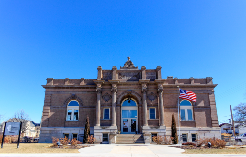 Historic brick building with a grand entrance, flanked by trees and an American flag, under a clear blue sky.