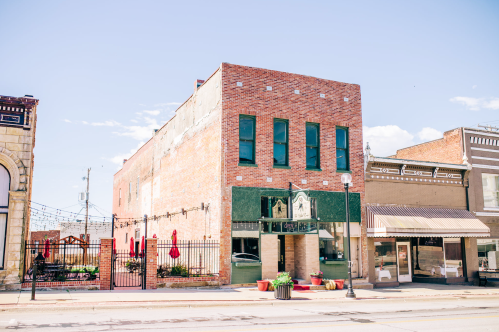 A brick building with green accents, featuring large windows and outdoor seating, set in a small town street.