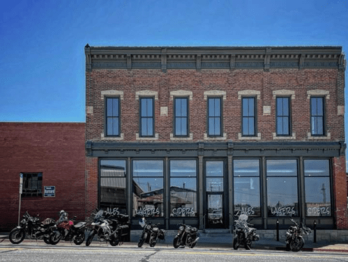 A brick building with large windows and a sign, parked motorcycles line the street in front under a clear blue sky.
