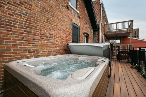 A hot tub on a wooden deck next to a brick wall, with a covered area and a balcony in the background.
