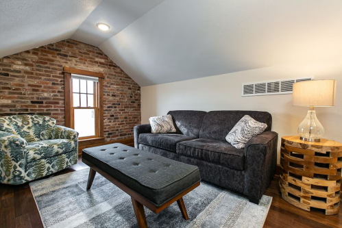 Cozy attic living space with a brick wall, a dark sofa, patterned chair, wooden table, and a stylish lamp.