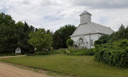 A white church with a steeple surrounded by greenery, set along a gravel road under a cloudy sky.