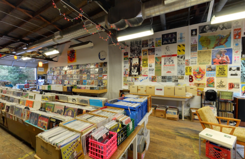 A cozy record store interior with shelves of vinyl records, colorful posters on the walls, and wooden flooring.