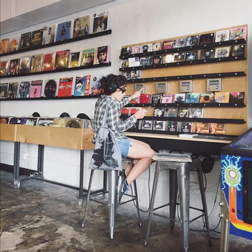 A person sits on a stool, browsing vinyl records in a stylish music store with colorful album covers on the wall.