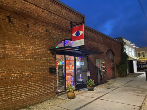 A brick building at night with an "Open" sign and a large eye logo above the entrance. Potted plants are outside.