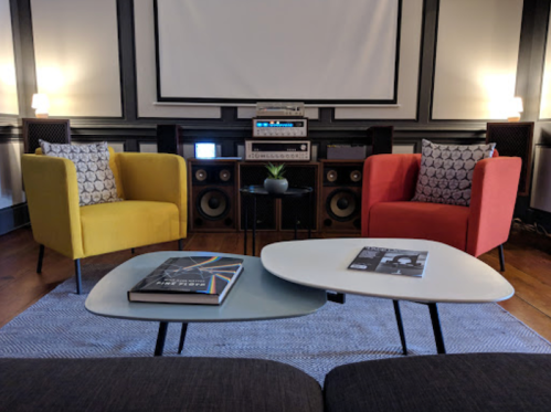 A cozy living room with two colorful chairs, a coffee table, and a sound system against a wall with a projector screen.