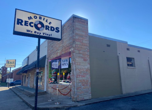 Exterior of Mobile Records store with a sign that reads "We Buy Vinyl" and a "Now Open" banner. Clear blue sky.