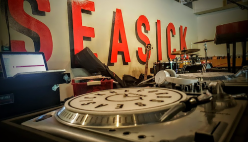 A DJ setup with turntables in front of a large "SEASICK" sign and musical instruments in the background.