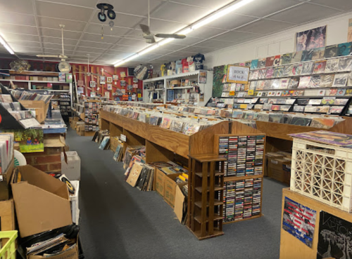 A cozy record store interior with rows of vinyl records, CDs, and wooden display tables. Boxes are stacked around the space.