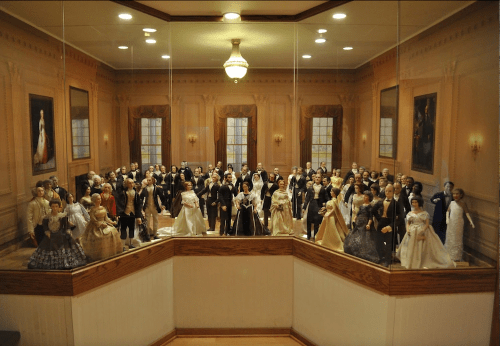 A display of numerous elegant dolls in formal attire, arranged in a room with vintage decor and large windows.