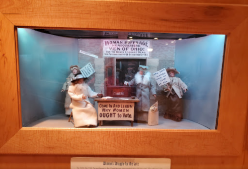 Display of figurines advocating for women's suffrage, with a sign reading "Come In and Learn Why Women Ought to Vote."