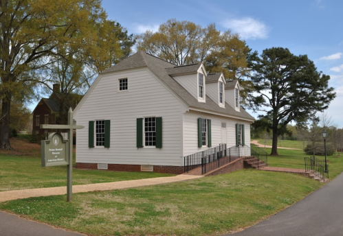 A historic white house with green shutters, surrounded by trees and a pathway, featuring a sign for a museum.