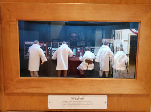A display of miniature figures in lab coats operating vintage control panels in a museum setting.