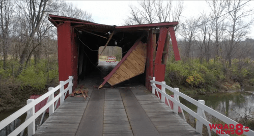 A damaged covered bridge with a collapsed roof, surrounded by trees and a river.