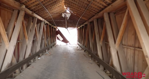 View inside a dilapidated covered bridge, with wooden beams and debris scattered on the floor, leading to bright light outside.