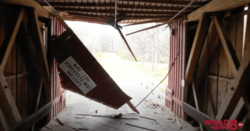 View from inside a damaged covered bridge, with debris and a caution sign visible.