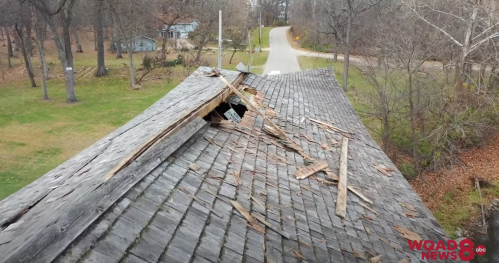 Aerial view of a damaged roof with missing shingles and a hole, surrounded by trees and a road in the background.