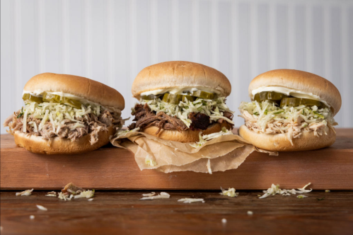 Three pulled pork sandwiches on a wooden board, topped with coleslaw and pickles, with some shredded cabbage scattered around.