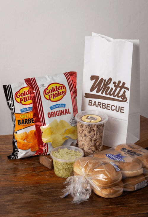 A table with bags of chips, a container of pulled pork, guacamole, hamburger buns, and a takeout bag labeled "Whitt's Barbecue."