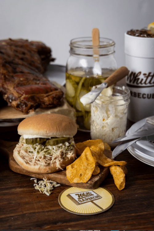 A pulled pork sandwich with pickles, coleslaw, and chips, alongside jars of pickles and a plate of ribs.