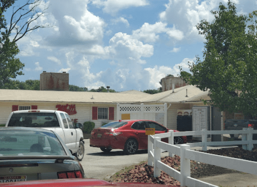 A red car and a white truck parked outside a restaurant with a white fence and cloudy sky in the background.