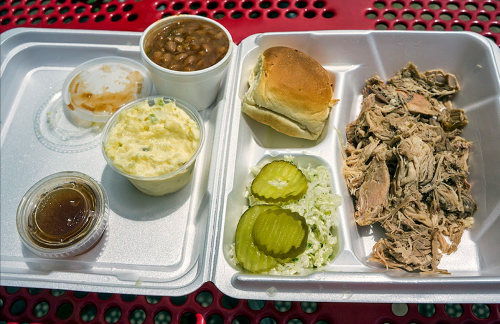 A takeout container with pulled pork, pickles, coleslaw, mashed potatoes, baked beans, and a roll.