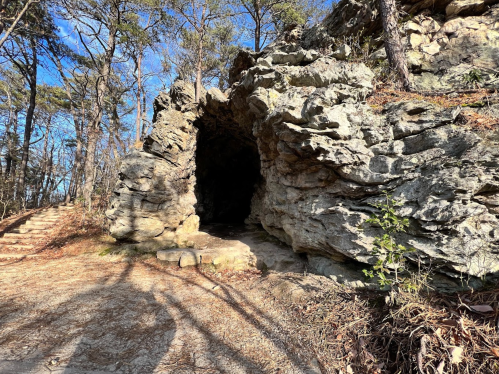 A rocky cave entrance surrounded by trees and a dirt path leading into the woods.