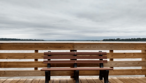 A wooden bench on a pier overlooking a calm river under a cloudy sky.