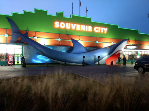 A large blue shark sculpture outside a brightly lit souvenir shop at dusk.